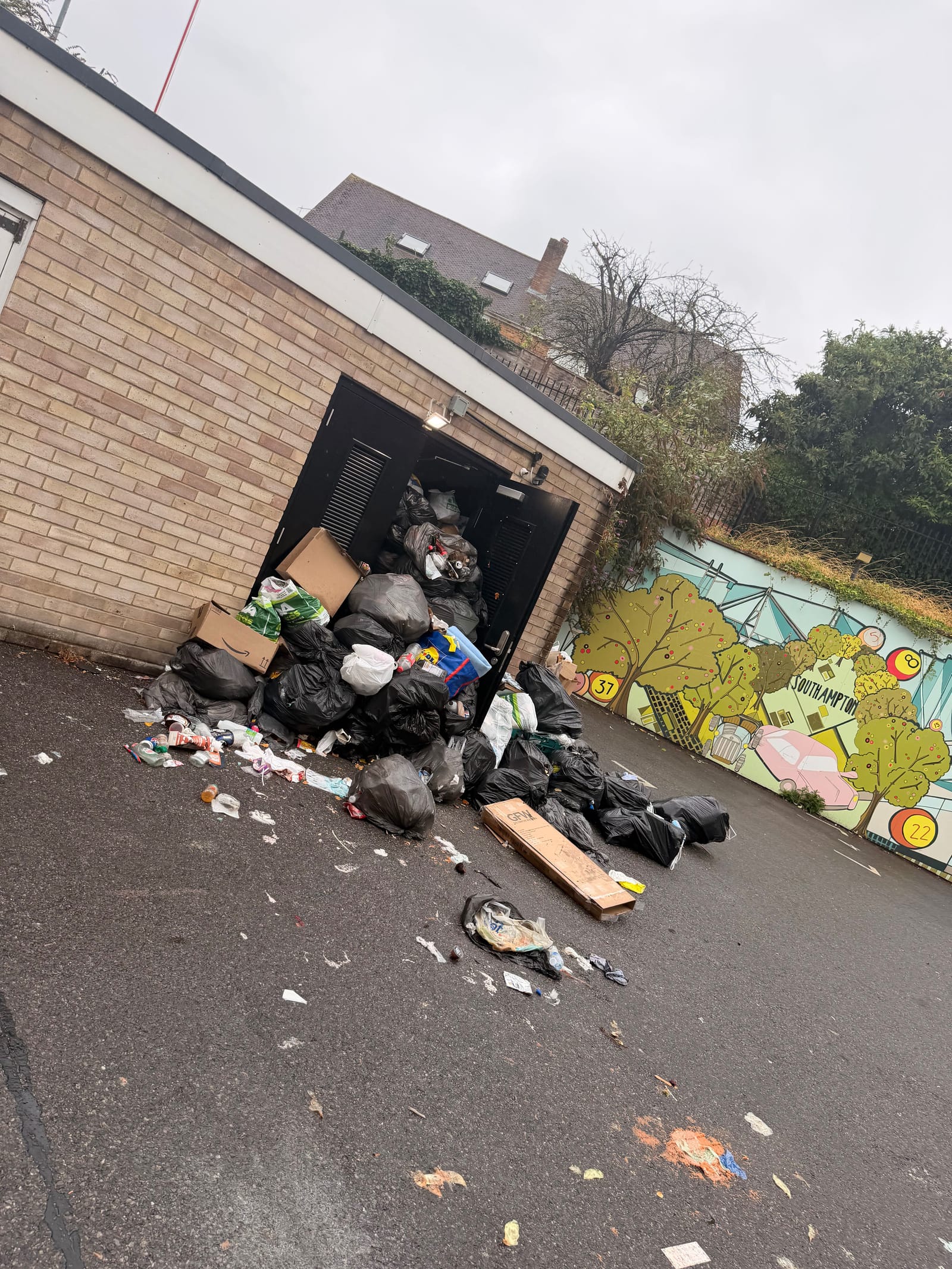 Bagged waste spilling out of a bin store doorway across a car park
